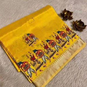 May include: Bright yellow fabric with a repeating pattern of dancing figures in colorful attire. The fabric has a gold border and is folded, with a soft, textured background. Two small, gold-colored lotus flower candle holders are in the background.
