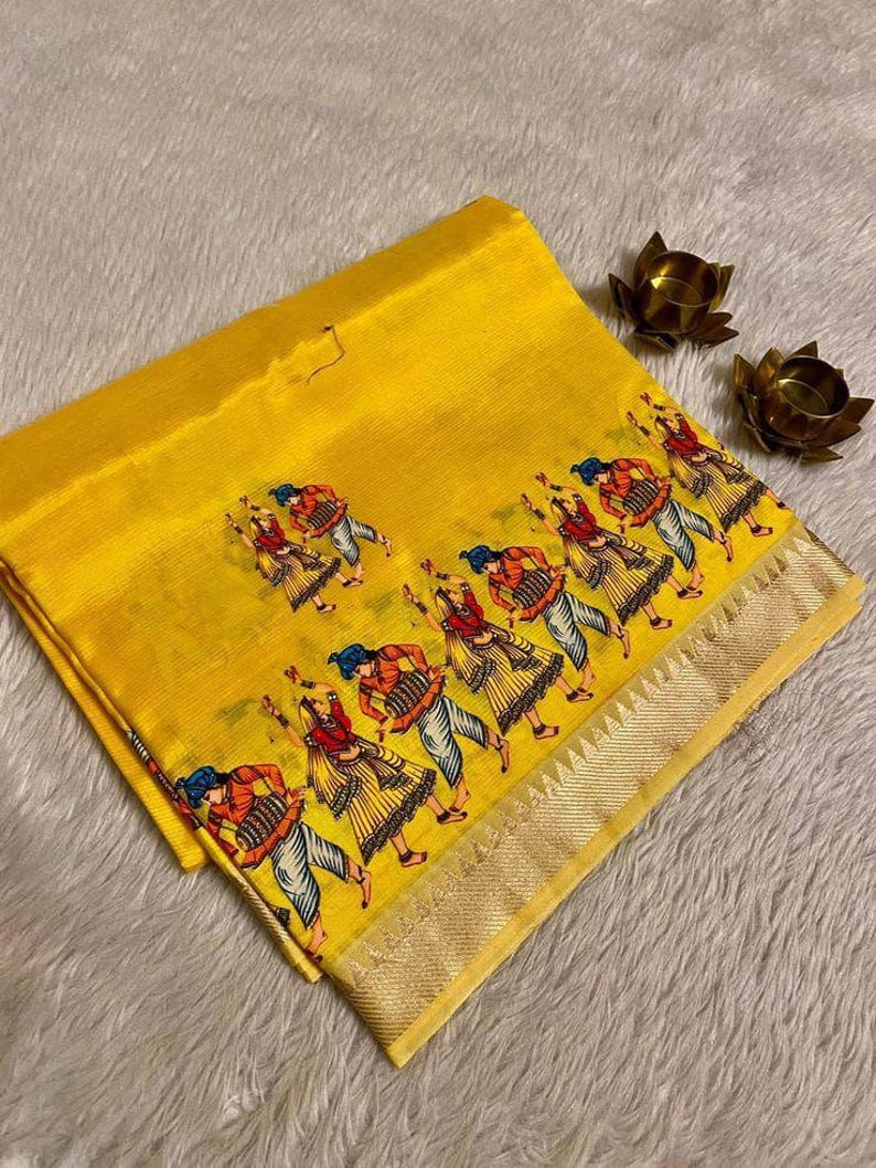 May include: Bright yellow fabric with a repeating pattern of dancing figures in colorful attire. The fabric has a gold border and is folded, with a soft, textured background. Two small, gold-colored lotus flower candle holders are in the background.