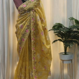 Peut inclure: Un sari jaune avec broderie florale, pr&eacute;sent&eacute; sur un mannequin. Le sari pr&eacute;sente des d&eacute;tails floraux roses et dor&eacute;s le long de la bordure et du corsage. Un collier de perles et un chemisier violet sont &eacute;galement visibles.