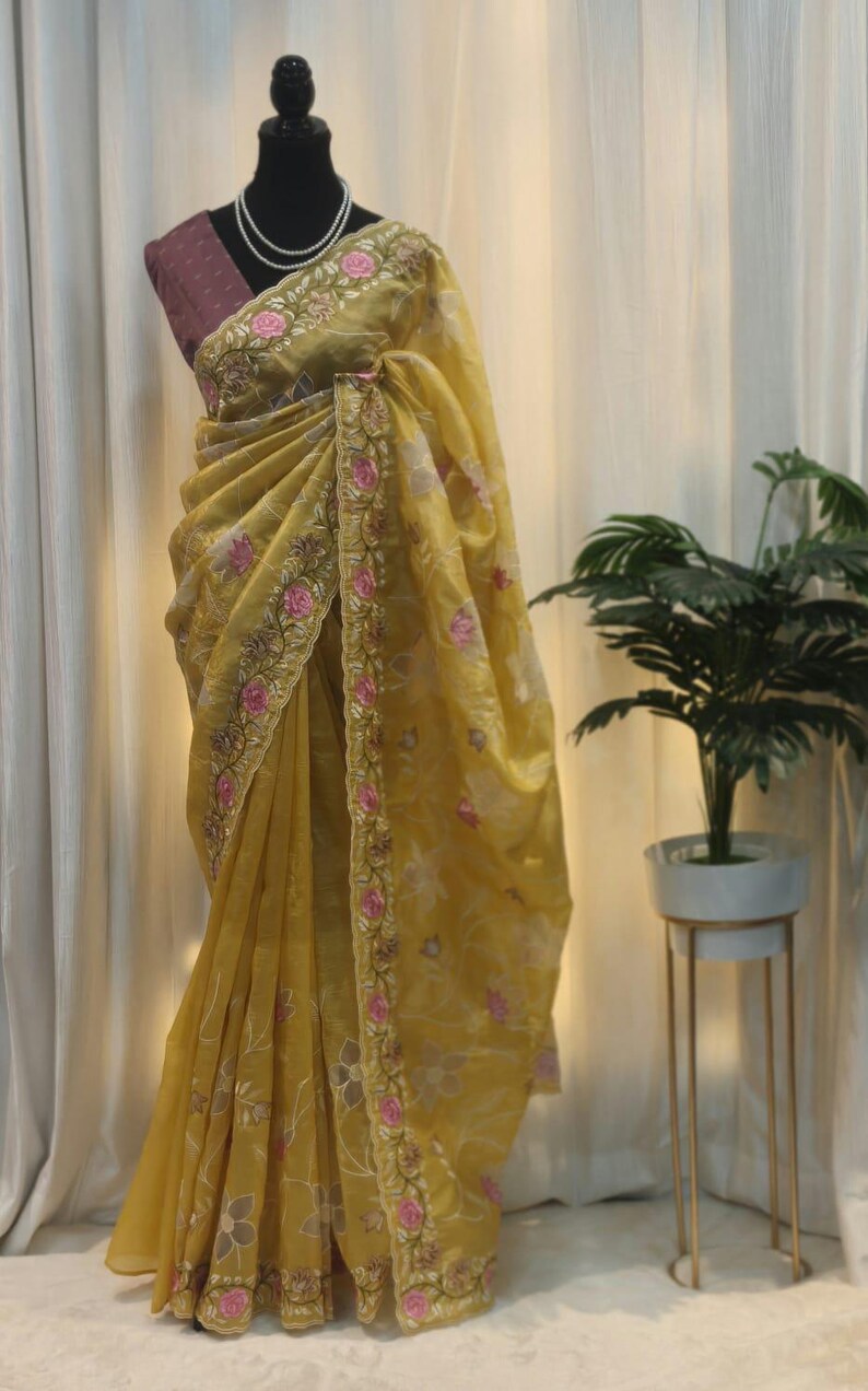 Peut inclure: Un sari jaune avec broderie florale, pr&eacute;sent&eacute; sur un mannequin. Le sari pr&eacute;sente des d&eacute;tails floraux roses et dor&eacute;s le long de la bordure et du corsage. Un collier de perles et un chemisier violet sont &eacute;galement visibles.