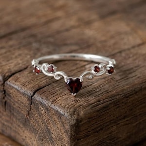 May include: A silver ring with a heart-shaped, deep red gemstone at the centre. The ring features a delicate, swirling design with smaller red gemstones. The ring is set against a rustic wooden surface.