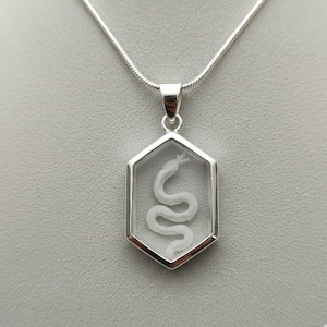 May include: A silver necklace featuring a hexagonal pendant with a clear center. Inside the pendant is a white, detailed snake. The necklace has a silver chain and clasp, set against a neutral background.