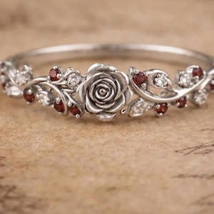Pode incluir: Um anel de tom prateado com um motivo central de rosa esculpida, ladeado por metal em forma de videira. O design é acentuado com pequenas pedras redondas de cor granada escura e pedras brancas transparentes ao longo da banda floral.