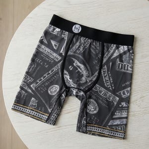 May include: Black boxer briefs with a repeating pattern of US $100 bills. The briefs have a black waistband with a white logo and gold chain detailing along the leg openings. The briefs are made of a soft, comfortable material.