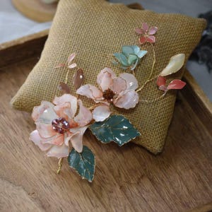 May include: A handcrafted floral brooch featuring delicate pink and white flowers with green leaves and gold wire accents. The brooch is adorned with small, sparkling crystals and rests on a burlap pillow, showcasing its intricate design.