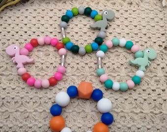 May include: Four colorful baby teething bracelets. Three bracelets feature round beads in shades of pink, green, blue, and white, with a dinosaur-shaped charm. The fourth bracelet has orange, white, and blue beads with a geometric center piece.