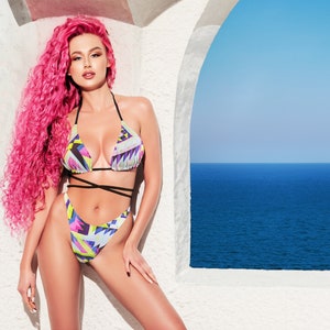 Puede incluir: Una mujer con el pelo rizado rosa lleva un bikini de estampado geométrico colorido. Está de pie frente a un arco blanco con vistas al mar.