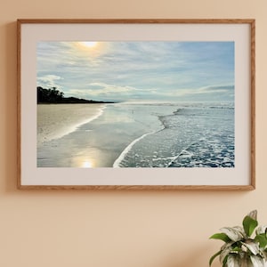 Puede incluir: Fotografía enmarcada de una escena de playa. La imagen captura una vista costera serena con una orilla arenosa, olas suaves y un cielo nublado. El sol es visible, reflejándose en la arena mojada y el agua. El marco es de madera marrón claro.
