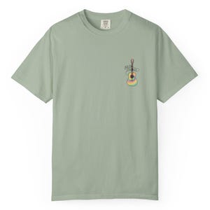 Op de afbeelding: Saliegroen t-shirt met een kleine, kleurrijke gitaarafbeelding op de voorkant. Het gitaarontwerp heeft regenboogkleuren en de tekst "Billy Strange" erboven. Het shirt heeft een ronde hals en korte mouwen.