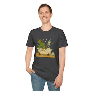 May include: Dark gray t-shirt featuring a whimsical illustration of green olives relaxing in a bathtub filled with bubbles. The olives have toothpicks and smiling faces. Bottles of gin are also in the image.