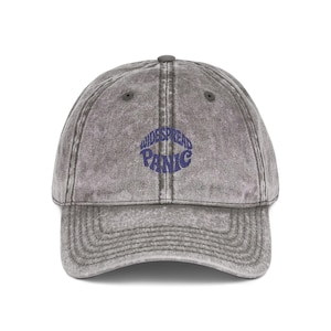 May include: A gray baseball cap with a curved brim and a rounded top. The cap features the text "WIDESPREAD PANIC" in a blue, stylized font across the front. The hat has a vintage, worn look.
