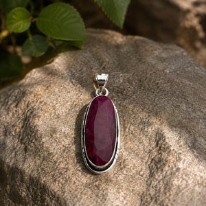 Può includere: Un ciondolo di forma ovale con una pietra preziosa sfaccettata rosso scuro incastonata in una cornice d'argento lucido. Il ciondolo include un gancio d'argento sulla parte superiore per l'attacco a una catena, con un bordo metallico liscio.