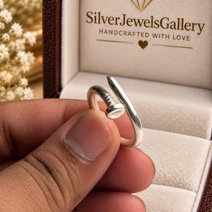 May include: A silver-toned ring shaped like a bent nail with a flat head and a textured shank. The ring is positioned in front of an open jewelry box featuring the text SilverJewelsGallery HANDCRAFTED WITH LOVE and a diamond icon.