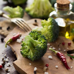 Pode incluir: Um close-up de floretes de br&oacute;colos frescos, pimentas vermelhas e gr&atilde;os de pimenta dispostos numa t&aacute;bua de cortar de madeira. Uma garrafa de azeite e um garfo est&atilde;o no fundo. Est&eacute;tica r&uacute;stica e natural.