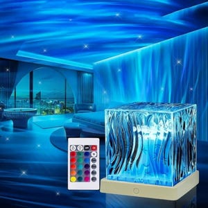 May include: A square-shaped water ripple projection lamp with a textured, translucent blue surface resting on a white base, accompanied by a rectangular remote control featuring multiple coloured buttons for light adjustment and settings.
