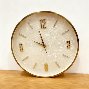 May include: A round wall clock with a gold-colored frame and hands. The clock face is white with a textured, mother-of-pearl effect. Gold numerals mark the 12, 3, 6, and 9 o'clock positions, with gold dashes for the other hour markers.