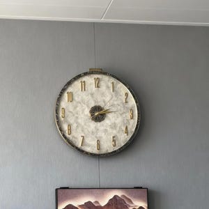 May include: A circular wall clock with a white face and gold numerals and hands. The clock has a gold frame and a decorative centre. The clock is hanging on a grey wall.