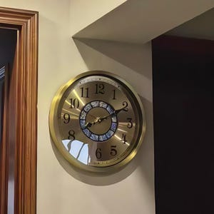 Könnte beinhalten: Eine runde, goldfarbene Wanduhr mit schwarzen Ziffern und Zeigern. Das Zifferblatt hat eine dekorative Mitte mit blauen und goldenen Akzenten. Die Uhr ist an einer beigefarbenen Wand befestigt.