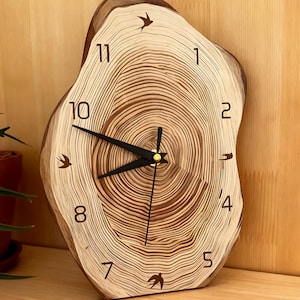 Puede incluir: Un reloj de pared de madera con una forma orgánica, que recuerda a una sección transversal de un tronco de árbol. La esfera muestra las vetas de la madera, con manecillas negras y detalles dorados. Siluetas de pájaros adornan la esfera. Dimensiones: aproximadamente 25 cm de diámetro.