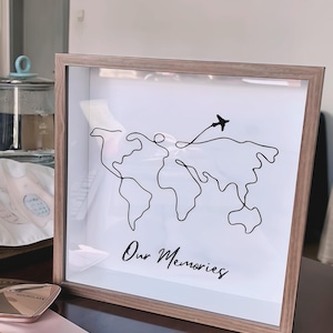 May include: A framed print featuring a minimalist world map design in black lines, with a small airplane and the words "Our Memories" in script. The frame is a light brown color.