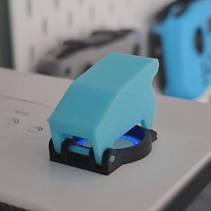 May include: A light blue 3D-printed switch cover with a black base. The switch cover has a unique shape and is designed to fit over a button. The background shows other electronic devices.