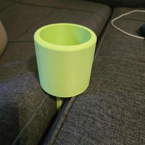 Puede incluir: Un recipiente cilíndrico verde claro con una parte superior redondeada y una base plana. El recipiente está sobre una superficie tapizada gris. El objeto parece ser una pequeña maceta o un soporte decorativo.