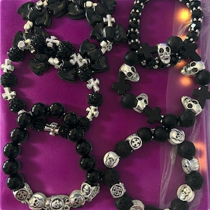 May include: Several black beaded bracelets with skull and cross designs. Some bracelets feature silver skulls and crosses, while others have black beads and accents. The bracelets are displayed on a purple surface.