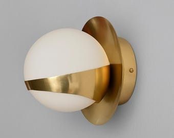 Puede incluir: Aplique de pared moderno con una luz de globo blanco esmerilado y una banda dorada, montado sobre un respaldo dorado en forma de pétalo. La luminaria está diseñada para montaje en pared, ofreciendo una solución de iluminación elegante.