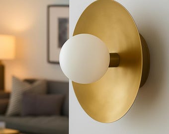 Puede incluir: Aplique de pared moderno con una gran placa trasera circular de latón cepillado. Una bombilla de globo de vidrio blanco y esférico se extiende desde el centro. El aplique está montado en una pared blanca, con una vista de un salón al fondo.