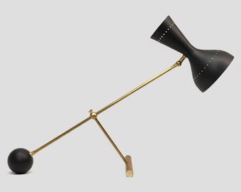 May include: A black and brass mid-century modern desk lamp. The lamp features a black, hourglass-shaped shade with small white dot details. The arm and base are brass-colored. The lamp has a round black base and a wooden support.