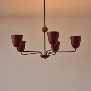 May include: A vintage-style chandelier featuring five burgundy lampshades. The light fitting has a dark metal frame, a wooden sphere, and a central rod. The chandelier is suspended from the ceiling.