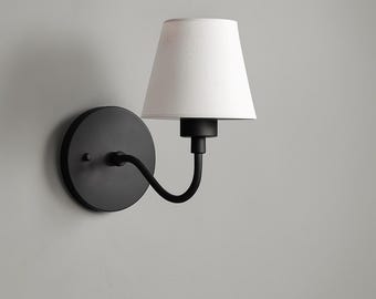 May include: A black wall sconce with a white lampshade. The sconce has a circular base and a curved arm that supports the lampshade. The lampshade is a classic tapered shape. The fixture is designed for indoor lighting.