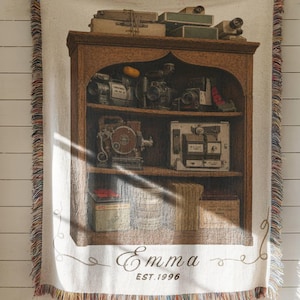 May include: A woven throw blanket featuring a brown wooden bookshelf graphic filled with vintage cameras and boxes. The text "Emma" and "EST. 1996" is printed in a script and serif font at the bottom. The blanket has a cream background and multicolored fringe edges.