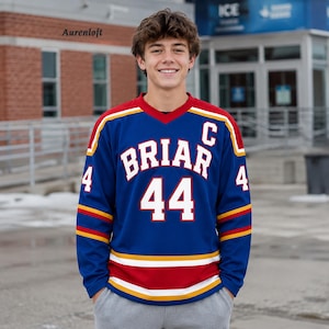Könnte beinhalten: Ein blaues Eishockeytrikot mit langen Ärmeln, das den Schriftzug BRIAR und die Nummer 44 in Weiß mit roten Umrandungen zeigt. Das Trikot hat ein weißes C auf der Brust, rote und gelbe Streifen an den Ärmeln und am Saum sowie die Nummer 4.