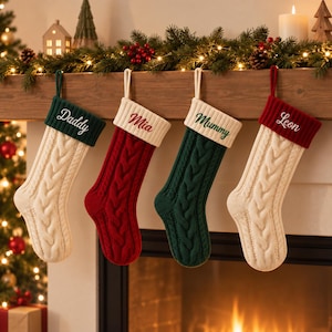 May include: Four cable-knit Christmas stockings hanging in a row. The first is cream with a dark green cuff embroidered with 'Daddy'. The second is red with a cream cuff embroidered with 'Mia'. The third is dark green with a cream cuff embroidered with 'Mummy'. The fourth is cream with a red cuff embroidered with 'Leon'.