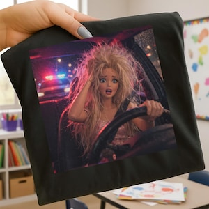 Puede incluir: Camiseta negra con una impresión de arte digital de una persona con aspecto de muñeca conduciendo un coche, con aspecto de shock. El fondo muestra luces de policía y una escena nocturna lluviosa. La persona sujeta el volante.