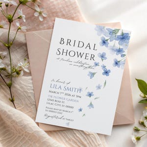 May include: A white bridal shower invitation with blue floral accents and the text "BRIDAL SHOWER". The invitation includes details for Lila Smith's shower, including the date, time, and location. A pink envelope is behind the invitation.