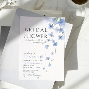 May include: A white bridal shower invitation with blue floral accents. The invitation includes the text "BRIDAL SHOWER" and event details, such as the date, time, and location. The invitation is registered at Target.