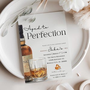 Peut inclure: Invitation d'anniversaire "Aged to Perfection" avec une bouteille de whisky et un verre de whisky avec des gla&ccedil;ons. L'invitation comprend le texte "Jake's 40th" et les d&eacute;tails de l'&eacute;v&eacute;nement. Le design est sur fond blanc.