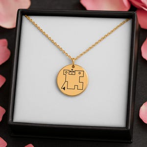May include: Gold-colored pendant necklace with a circular charm featuring a black geometric design. The necklace is presented in a black box with a white interior, and is surrounded by pink rose petals.