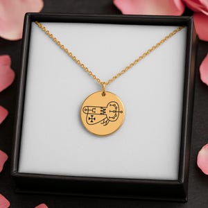 Op de afbeelding: Goudkleurige hangerketting met een cirkelvormige bedel met een zwart grafisch ontwerp. De ketting wordt gepresenteerd in een zwarte doos met een witte binnenkant en de ketting is delicaat. De achtergrond bevat roze bloemblaadjes.