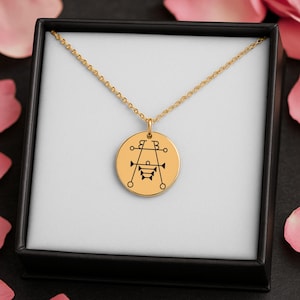 May include: A gold-colored pendant necklace with a circular charm featuring a black graphic design. The necklace is displayed in a black box with a white interior, surrounded by pink flower petals.