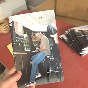 Könnte beinhalten: Ein Foto eines Mannes, der an einem Spielautomaten sitzt. Der Mann trägt ein graues Hemd, blaue Jeans und Turnschuhe. Der Spielautomat hat einen dunklen Schrank mit beleuchteten Knöpfen und Bildschirmen. Das Foto wird von einer Hand auf einer roten Oberfläche gehalten.