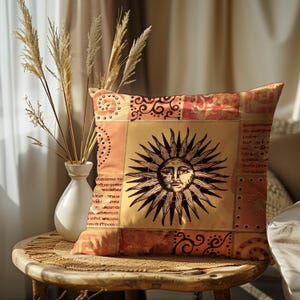 May include: Decorative throw pillow with a sun design. The pillow features a central sun graphic with a face, surrounded by a patchwork of orange, peach, and tan fabrics. The pillow is on a wooden table with a vase of dried plants.