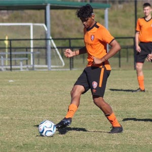 Puede incluir: Un jugador de fútbol con camiseta naranja y pantalones cortos negros patea un balón en un campo verde. El jugador lleva calcetines naranjas y botas negras. Otro jugador está en el fondo.
