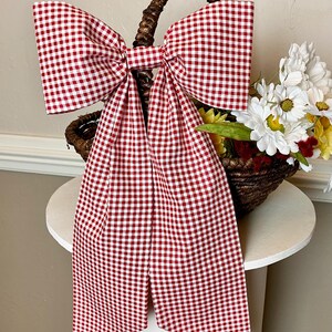 May include: A large red and white gingham bow adorns a woven basket filled with flowers. The bow is the focal point, with long, flowing tails. The basket sits on a white, round table, creating a charming, rustic aesthetic.