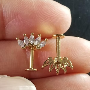 May include: Two gold-colored, decorative ear studs. One stud features a crown of clear, marquise-cut stones. The other stud has a floral design on the back. Both studs have a post and flat disc backing.