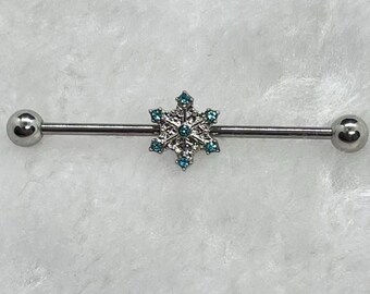May include: A silver industrial barbell earring featuring a snowflake design. The snowflake is embellished with small, light blue gemstones. The barbell has a ball at each end.