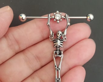 May include: A silver industrial barbell earring with a skeleton charm. The barbell has a small, clear gem in the centre and a ball on each end. The skeleton is holding the barbell above its head.