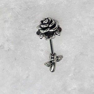 Puede incluir: Un piercing plateado con un diseño de rosa. La rosa está detallada con múltiples pétalos y se asienta sobre un poste recto. Pequeñas hojas están presentes cerca de la base del poste. La joya está sobre un fondo blanco texturizado.